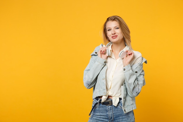 Portrait of cheerful pretty young woman in denim casual clothes with headphones looking camera isolated on bright yellow orange wall background in studio. People lifestyle concept. Mock up copy space.