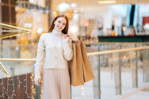 Portrait of cheerful pretty young woman holding shopping bags with purchases on shoulder and smiling looking at camera in mall center