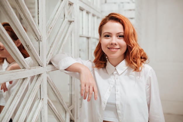 Portrait of cheerful redhead young woman dressed in white shirt standing near mirror in cafe