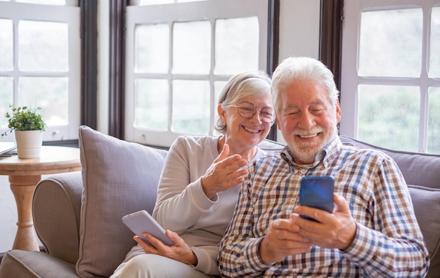 Portrait of cheerful senior couple while sitting together on sofa laughing Elderly happy couple relaxing while using smartphones