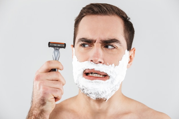 Portrait of a cheerful shirtless bearded man standing isolated over white, face covered with shaving foam, holding a razor