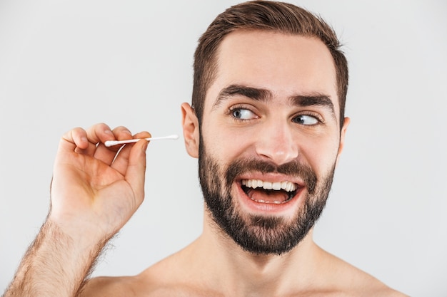 Portrait of a cheerful shirtless bearded man standing isolated over white, using cotton swab