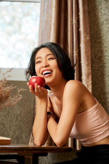 Портрет веселой стройной молодой женщины, едящей вкусное сладкое красное яблоко в качестве закуски
