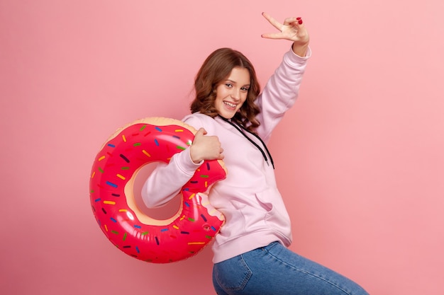 Portrait of cheerful smiling curly haired teenage girl in hoodie holding pink donut rubber ring and showing victory sign on fingers rest Indoor studio shot isolated on pink background