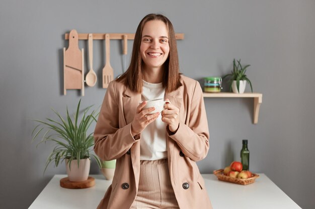 Portrait of cheerful smiling delighted young adult woman wearing beige suit standing near table on kitchen at home drinking coffee in the morning looking at camera