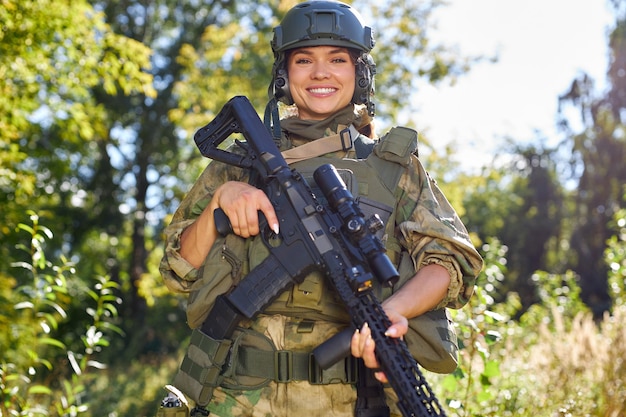 Portrait of cheerful smiling female soldier with rifle gun in hands in green military suit and hat