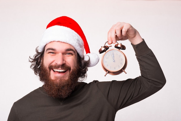 Portrait of cheerful smiling man with beard wearing santa claus hat and holding alarm clock