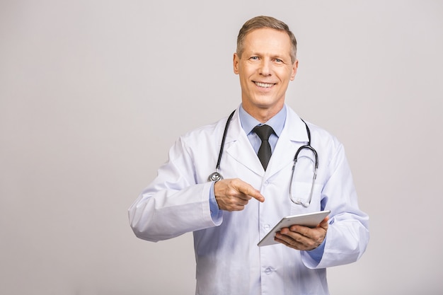 Портрет жизнерадостного усмехаясь старшего врача с стетоскопом используя таблетку ПК изолированную на серой стене.