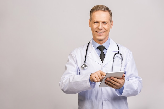 Portrait of a cheerful smiling senior medical doctor with stethoscope using pc tablet isolated on grey wall.