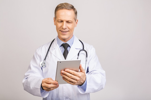 Portrait of a cheerful smiling senior medical doctor with stethoscope using pc tablet isolated on grey wall.