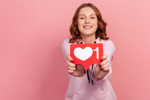 Portrait of cheerful smiling teenage girl showing heart icon at camera, recommending to follow, like me sign. Indoor studio shot isolated on pink background