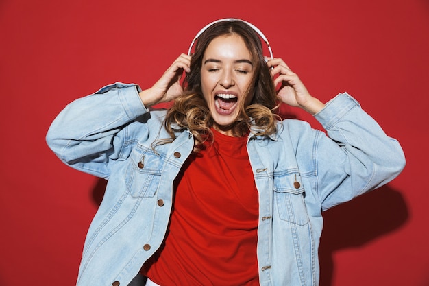 Portrait of a cheerful stylish young woman wearing denim jacket standing isolated over red wall, listening to music with headphones, dancing