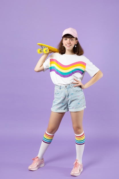 Portrait of cheerful teen girl in vivid clothes standing, holding yellow skateboard isolated on violet pastel wall background in studio. People sincere emotions, lifestyle concept. Mock up copy space.