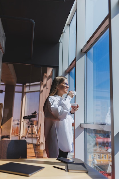 Portrait of a cheerful woman in classic glasses smiling in her free time in a cafe with coffee positive jewish woman in a white shirt desk with laptop remote work