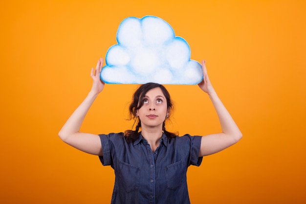 Portrait of cheerful woman holding and looking up on a thought cloud in studio over yellow background. Copy space available. Girl with placard. GIrl with announcement card.