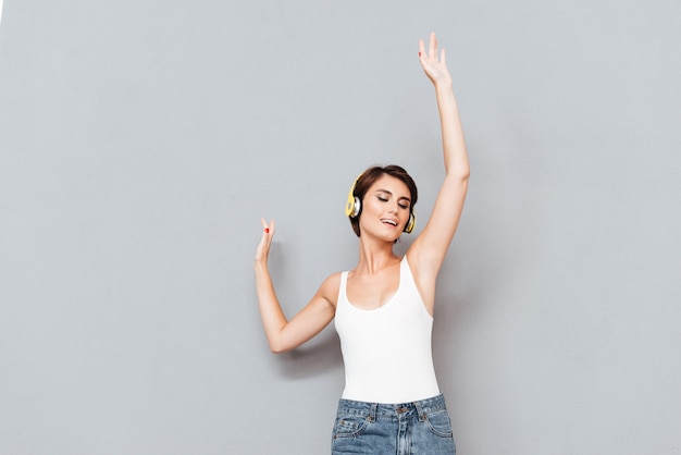 Portrait of a cheerful woman listening music in headphones and singing with raised hands up isolated on a gray background