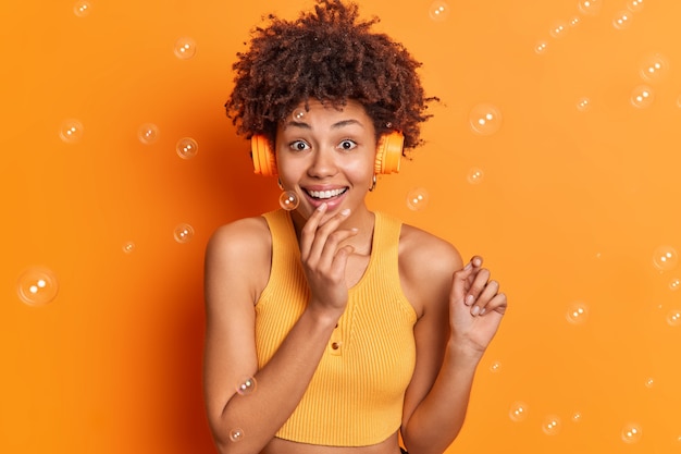 Portrait of cheerful woman looks gladfully at camera has natural curly hair listens audio track via headphones dressed casually isolated on vivid orange wall with soap bubbles around