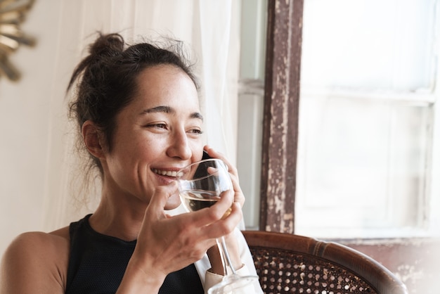 Portrait of cheerful woman in summer clothes talking on smartphone and drinking wine while sitting on armchair in apartment