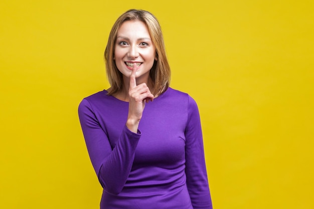Portrait of cheerful woman in tight purple dress making silence quiet gesture with finger on her mouth, asking to keep secret gossip. indoor studio shot isolated on yellow background