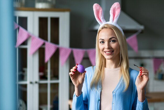 Portrait of cheerful woman wearing in bunny eggs and standing with a brush and violet egg in her hands. 