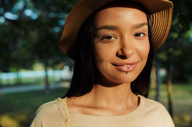 Portrait of cheerful woman wearing lip piercing and straw hat looking at camera in green park