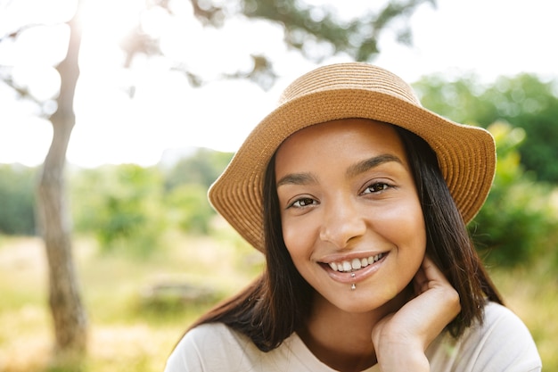 Портрет веселой женщины в соломенной шляпе и пирсинге губ, улыбающейся в камеру во время прогулки в зеленом парке