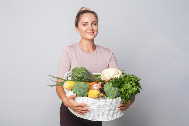 Portrait of cheerful woman with hair bun holding basket of fresh vegetables and looking at camera, vegetarian diet, healthy food, copy space for advertising. indoor studio shot, grey background