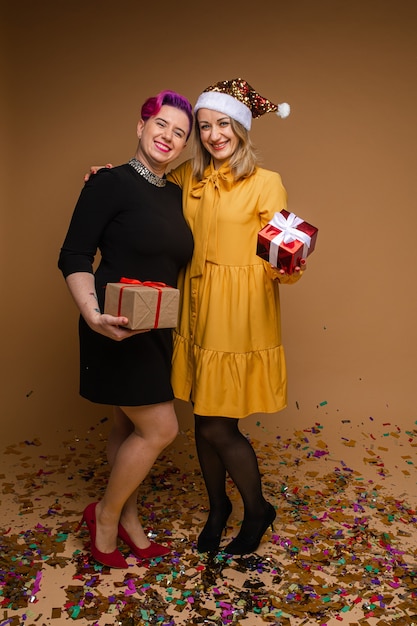 Photo portrait of cheerful women exchanging christmas gifts and smiling. blonde wearing sparkling santa hat and yellow dress