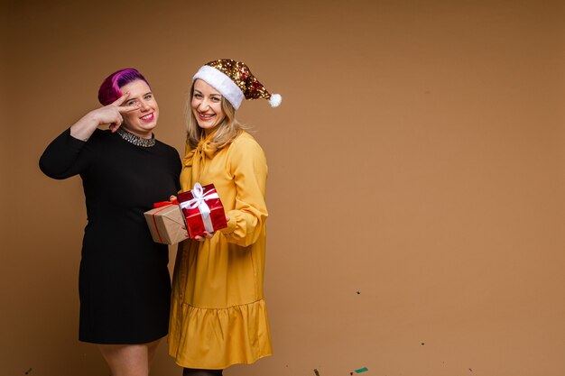 Portrait of cheerful women exchanging Christmas gifts and smiling. Blonde wearing sparkling Santa hat and yellow dress