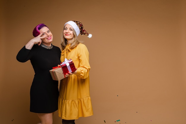 Portrait of cheerful women exchanging Christmas gifts and smiling. Blonde wearing sparkling Santa hat and yellow dress