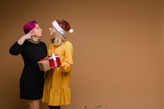 Portrait of cheerful women exchanging Christmas gifts and smiling. Blonde wearing sparkling Santa hat and yellow dress