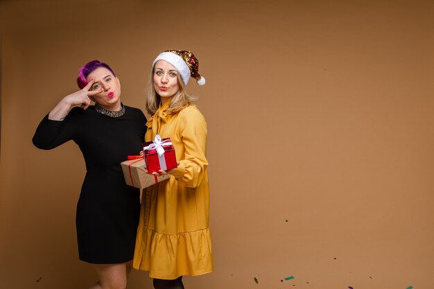 Portrait of cheerful women exchanging Christmas gifts and smiling. Blonde wearing sparkling Santa hat and yellow dress