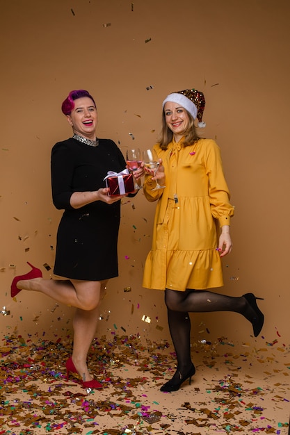 Portrait of cheerful women exchanging Christmas gifts and smiling. Blonde wearing sparkling Santa hat and yellow dress