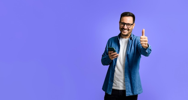 Portrait of cheerful young adult man in denim shirt using smart phone and showing thumbs up sign while standing isolated over blue background