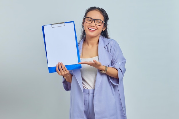 Portrait of cheerful young Asian business woman showing blank clipboard with palms isolated on purple background