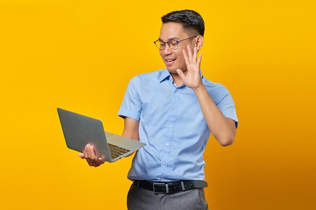 Portrait of cheerful young asian man Asian in glasses using a laptop doing a video call gesturing hi to friends isolated on yellow background businessman and entrepreneur concept