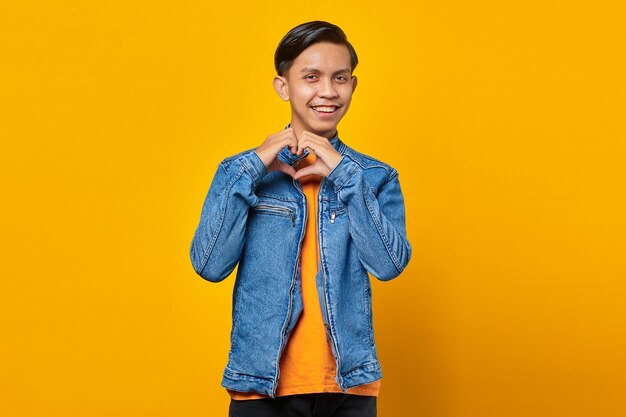 Portrait of cheerful young Asian man making heart sign with fingers on yellow background