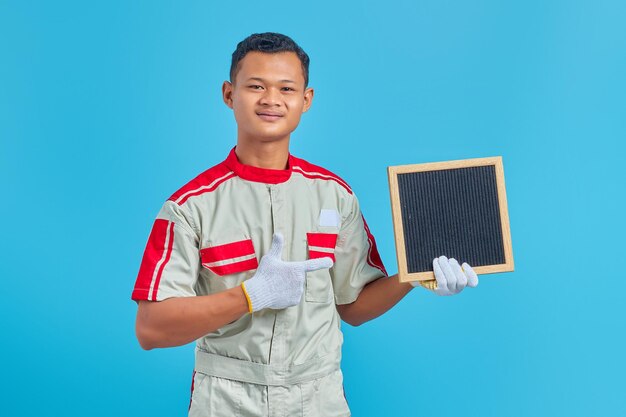 Portrait of cheerful young asian mechanic pointing at blank board with finger over blue background
