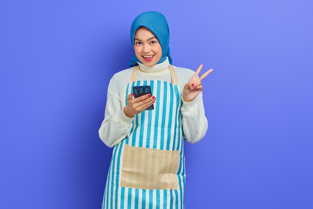 Портрет веселой молодой азиатки-мусульманки 20 лет в хиджабе и фартуке держит мобильный телефон, показывая знак мира пальцами, изолированными на фиолетовом фоне. Концепция мусульманского образа жизни домохозяйки