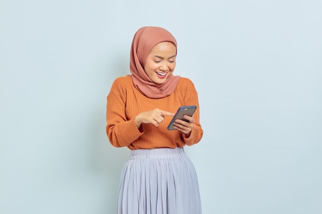 Portrait of cheerful young asian muslim woman in brown sweater\
and hijab using mobile phone isolated on white background muslim\
lifestyle concept