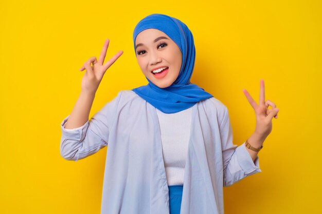 Portrait of cheerful young asian muslim woman showing peace
gesture isolated over yellow background