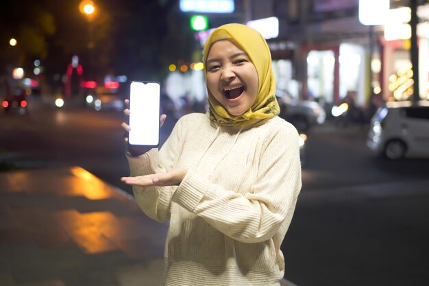 Portrait of cheerful young Asian Muslim woman in white sweater and hijab pointing to blank mobile phone screen in night City Street Full of Neon Light urban style