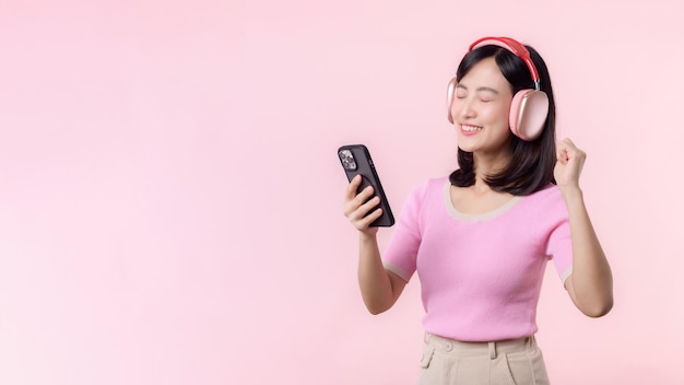 肖像画の陽気な若いアジア女性は、ピンクに対してスマートフォンの音楽アプリケーションでオーディオを聴くことを楽しみます