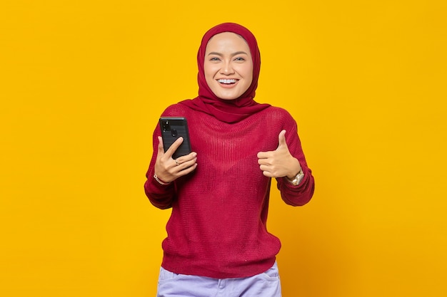 Portrait of cheerful young Asian woman holding mobile phone and showing thumbs up sign isolated on yellow background