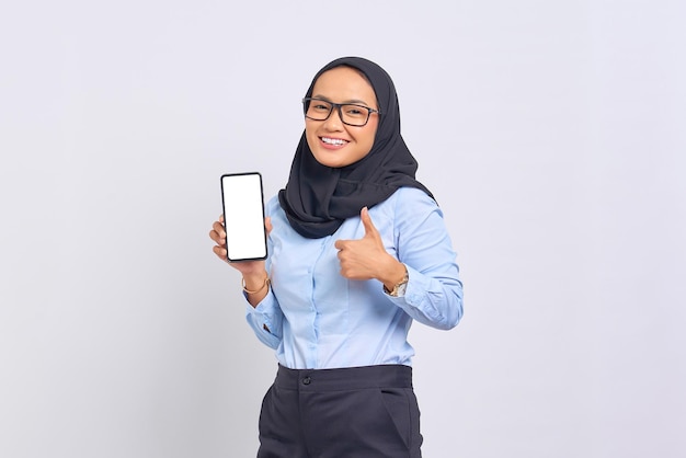 Portrait of cheerful young Asian woman showing blank screen mobile phone and gesturing thumbs up isolated on white background