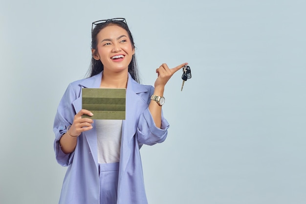 Portrait of cheerful young Asian woman showing vehicle book and holding vehicle keys isolated on white background