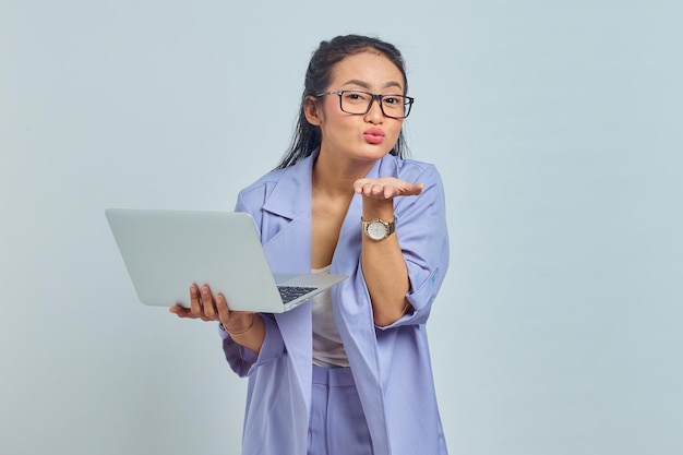 Portrait of cheerful young Asian woman standing using laptop and sending forward air kiss isolated over white background