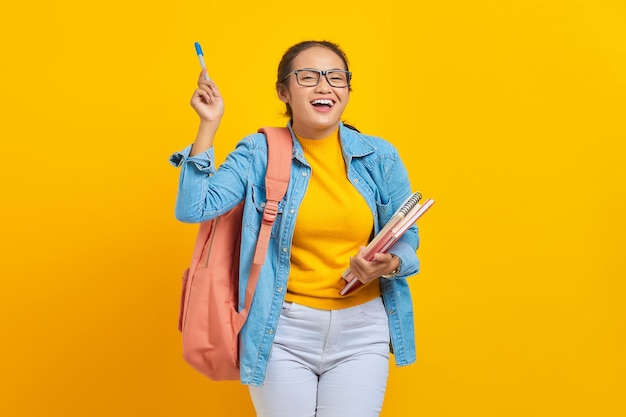 배낭을 메고 책을 들고 노란색 배경에 격리된 펜으로 복사 공간을 가리키는 캐주얼 옷을 입은 쾌활한 젊은 아시아 여성 학생의 초상화 대학 대학 개념 교육