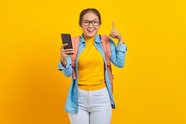 Portrait of  cheerful young Asian woman student in casual clothes with backpack holding mobile phone and pointing up space with fingers isolated on yellow background