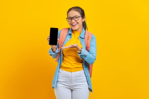 黄色の背景で隔離の手のひらで空白の画面の携帯電話を指すバックパックとカジュアルな服装で陽気な若いアジアの女性学生の肖像画。大学の概念における教育